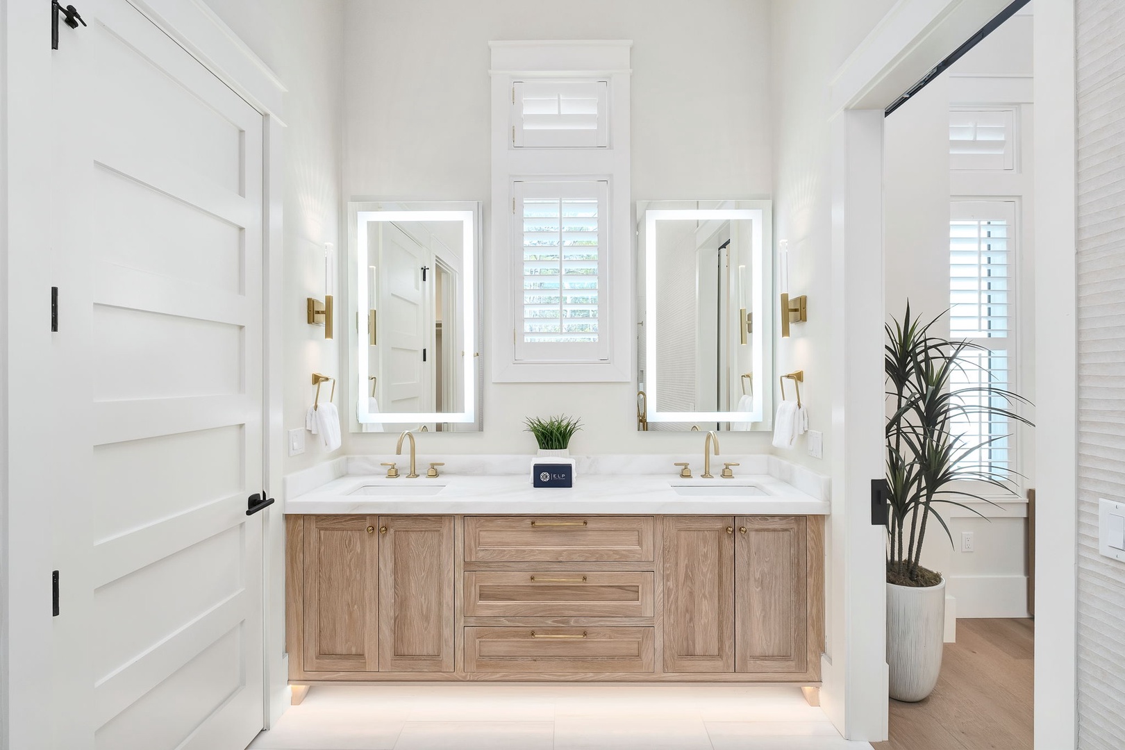 Step into your private sanctuary with a beautifully designed dual-vanity bathroom featuring warm wood cabinetry, elegant gold fixtures, and bright natural light filtering through plantation shutters—your perfect place to unwind.