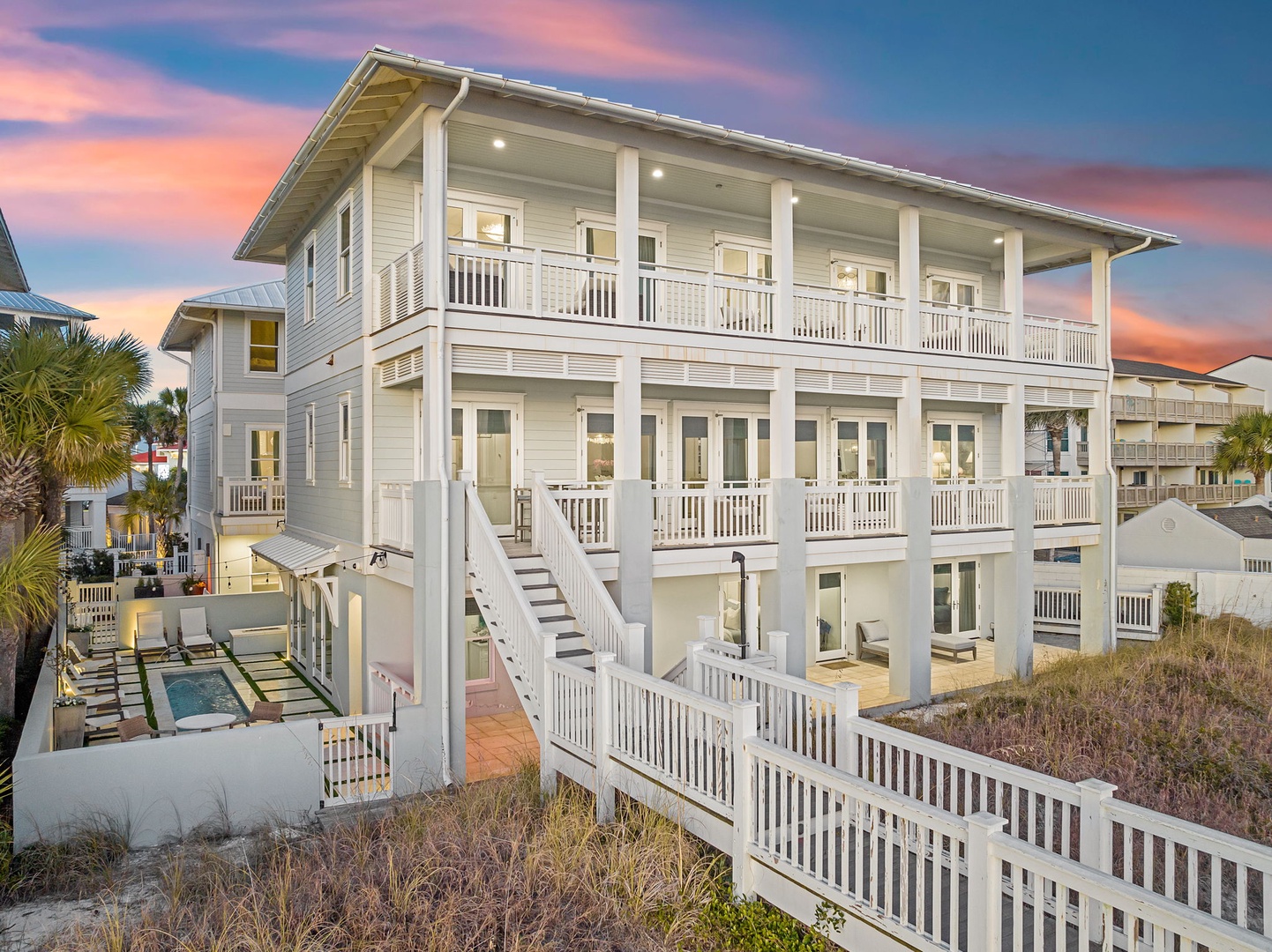 Elegant three-story beachfront property featuring expansive covered porches, coastal architecture, and a private pool area, situated among dunes with vibrant sunset skies overhead.