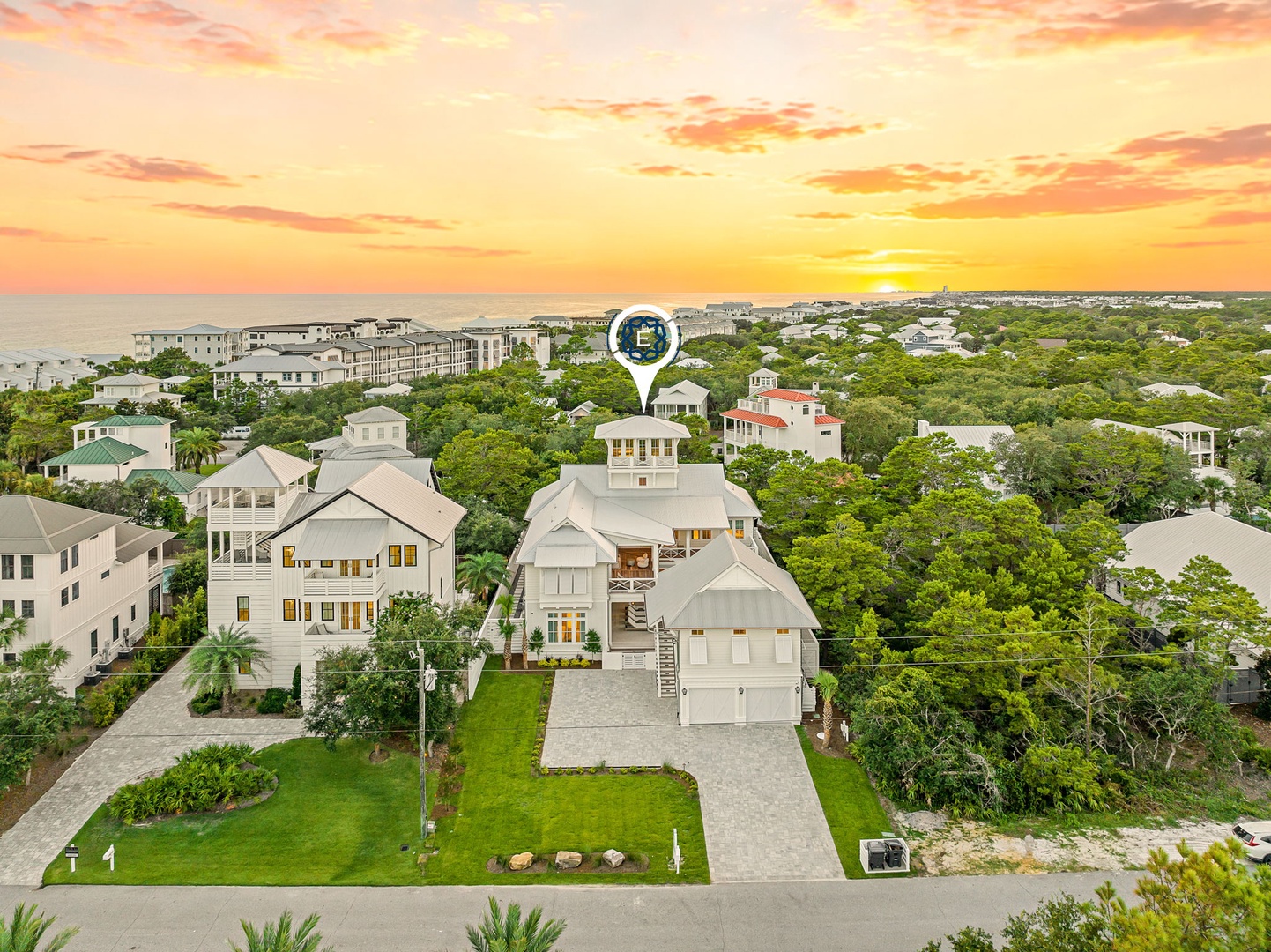 Aerial view of a coastal vacation rental marked with a property marker, nestled in a lush residential area with the ocean visible in the distance at sunset.