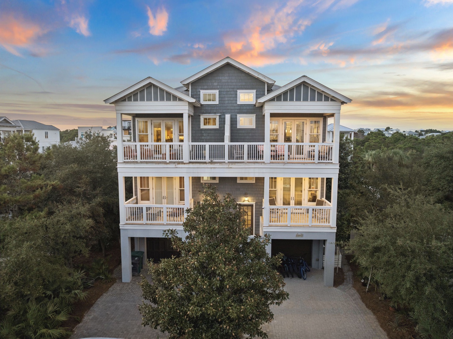 A striking three-story coastal home with twin covered porches set against peaceful natural surroundings, captured at dusk as warm interior lights begin to glow.