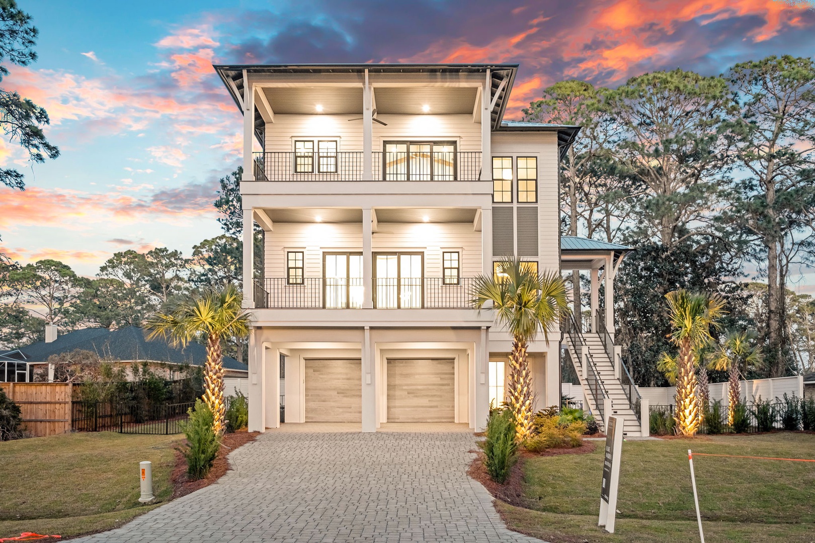 A stunning three-story coastal home with elegant balconies, surrounded by mature trees and palm trees under a vibrant sunset sky.