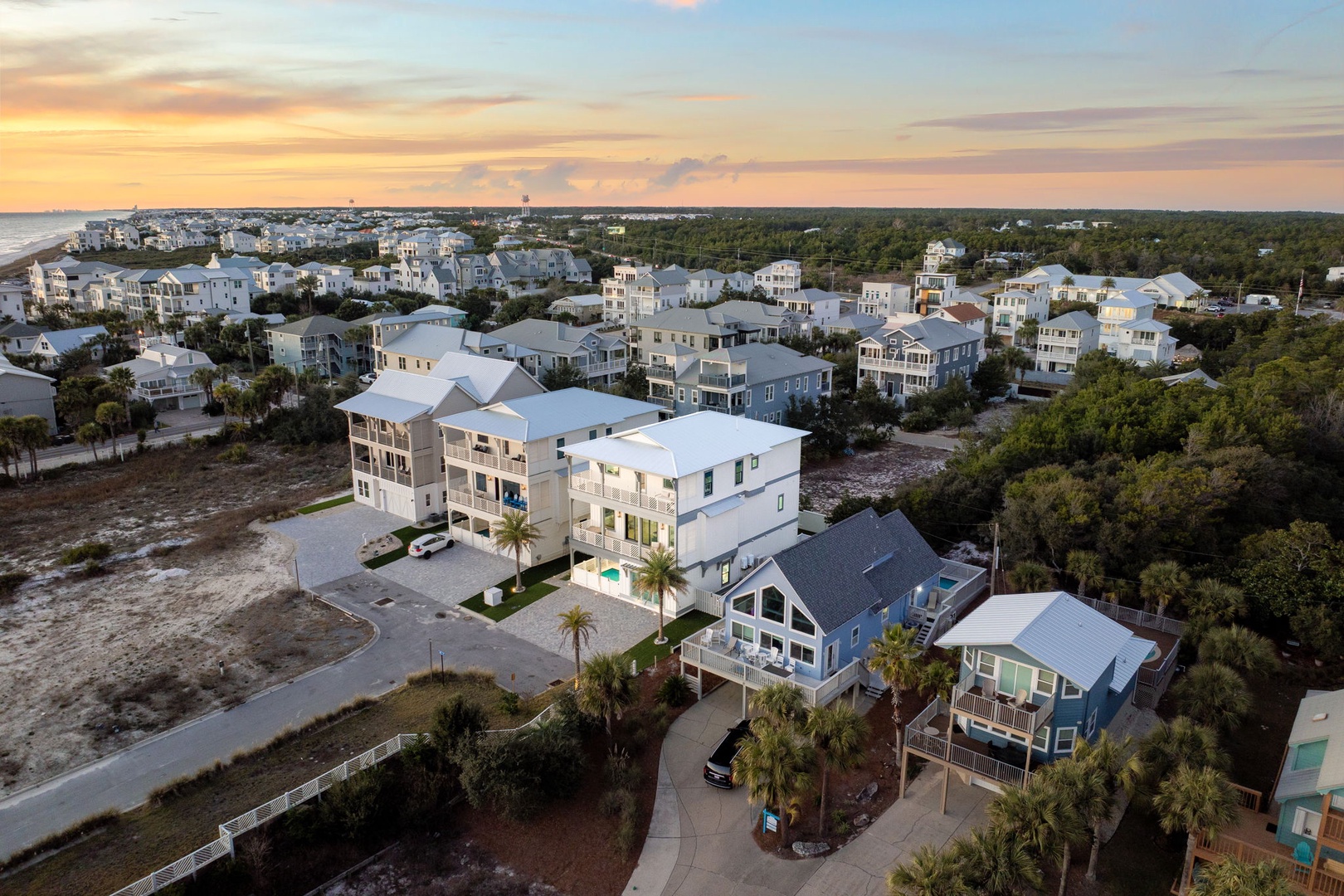 Aerial view of a coastal community featuring modern beach homes surrounded by lush vegetation and sandy terrain at sunset.