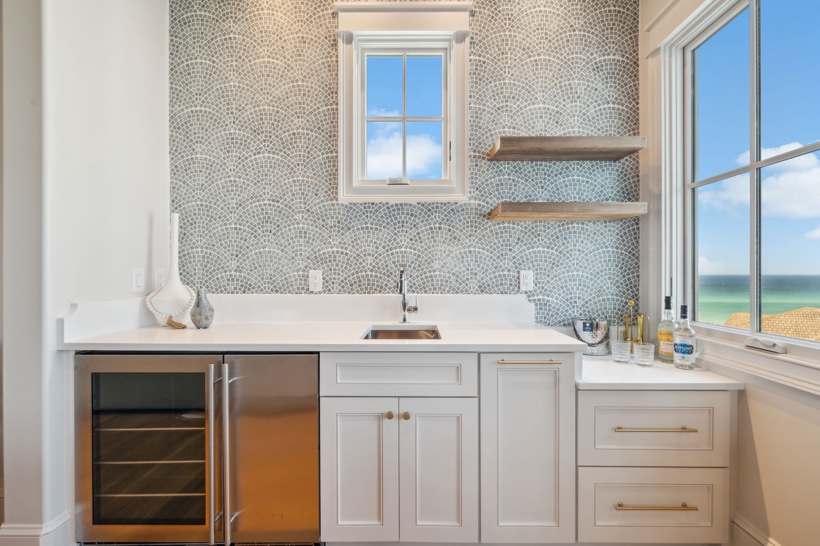 Your stylish wet bar features elegant mosaic tilework, white cabinetry, and a wine fridge—perfect for entertaining while enjoying coastal views through large windows.