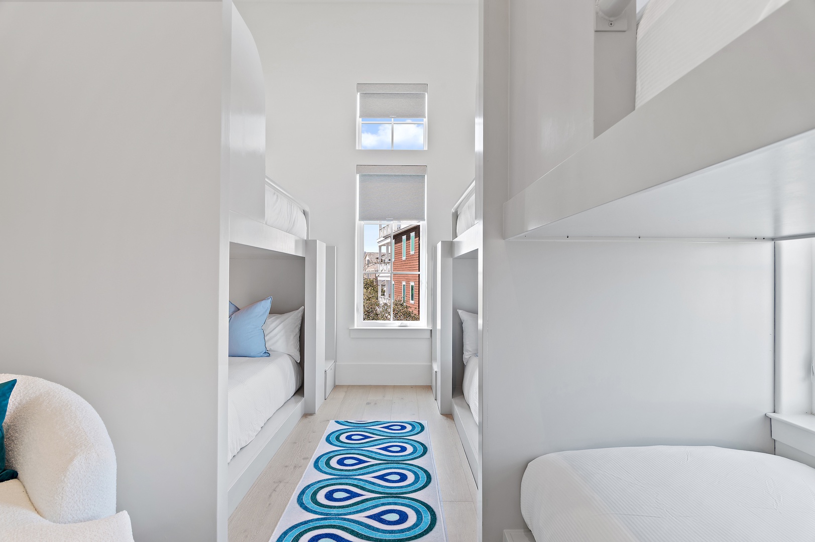 Crisp white built-in bunks line the walls, creating your perfect family retreat. Sunlight streams through tall windows, while a cheerful blue rug adds warmth to the bright, modern space.