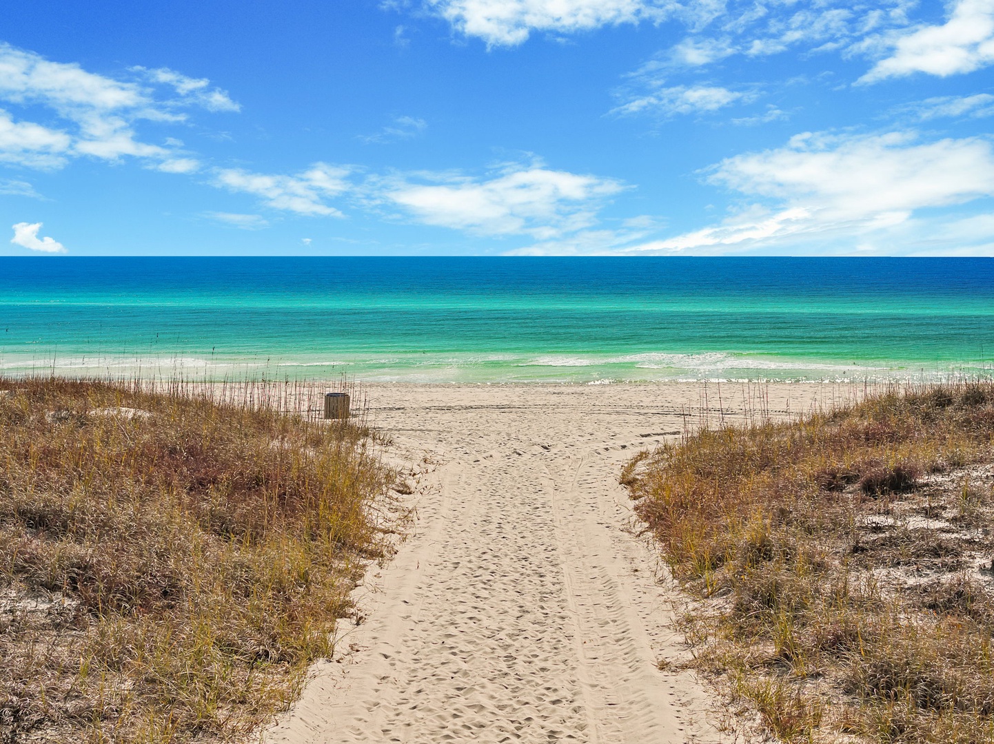 A sandy path leads through coastal dunes to pristine turquoise waters and white sand beach under brilliant blue skies.
