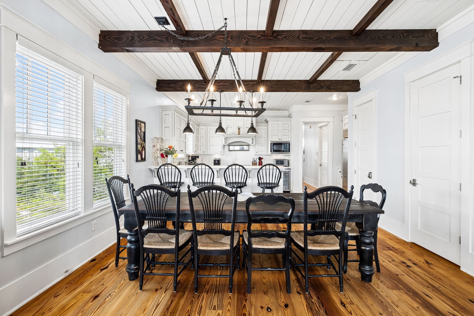 You'll gather around this stunning farmhouse table under rustic wooden beams, with natural light pouring through oversized windows and the bright, open kitchen just steps away—perfect for memorable meals with loved ones.