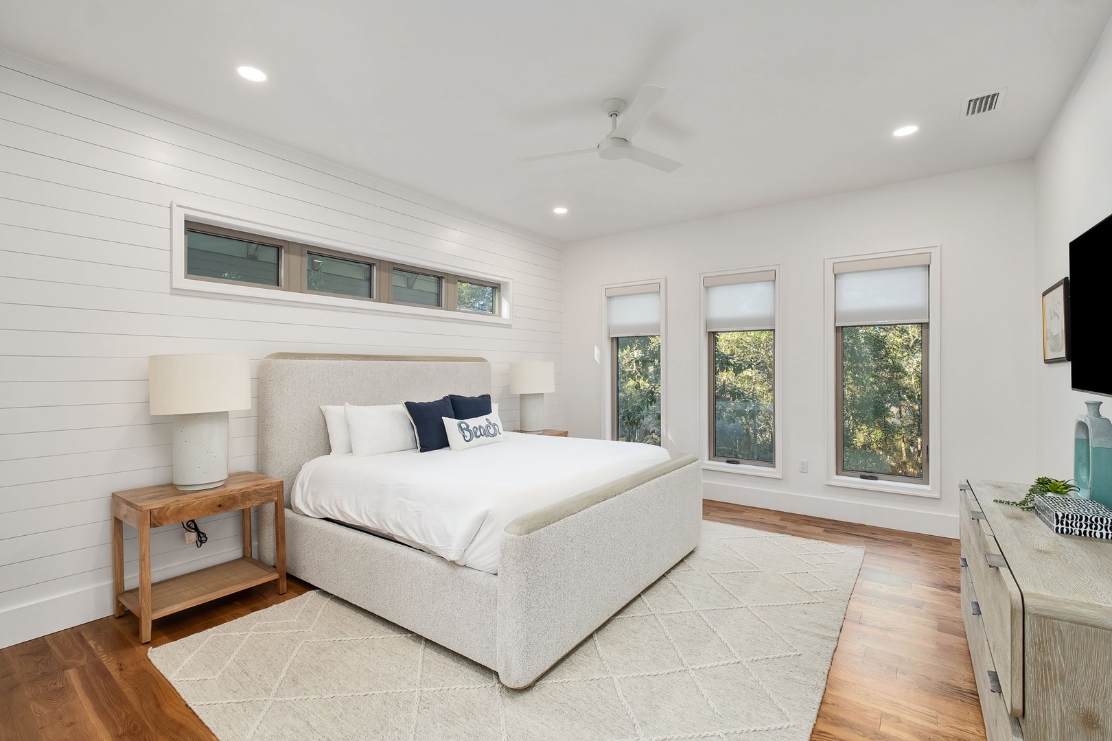 Drift off in your serene bedroom featuring crisp white linens, warm wood floors, and natural light streaming through windows overlooking lush greenery.