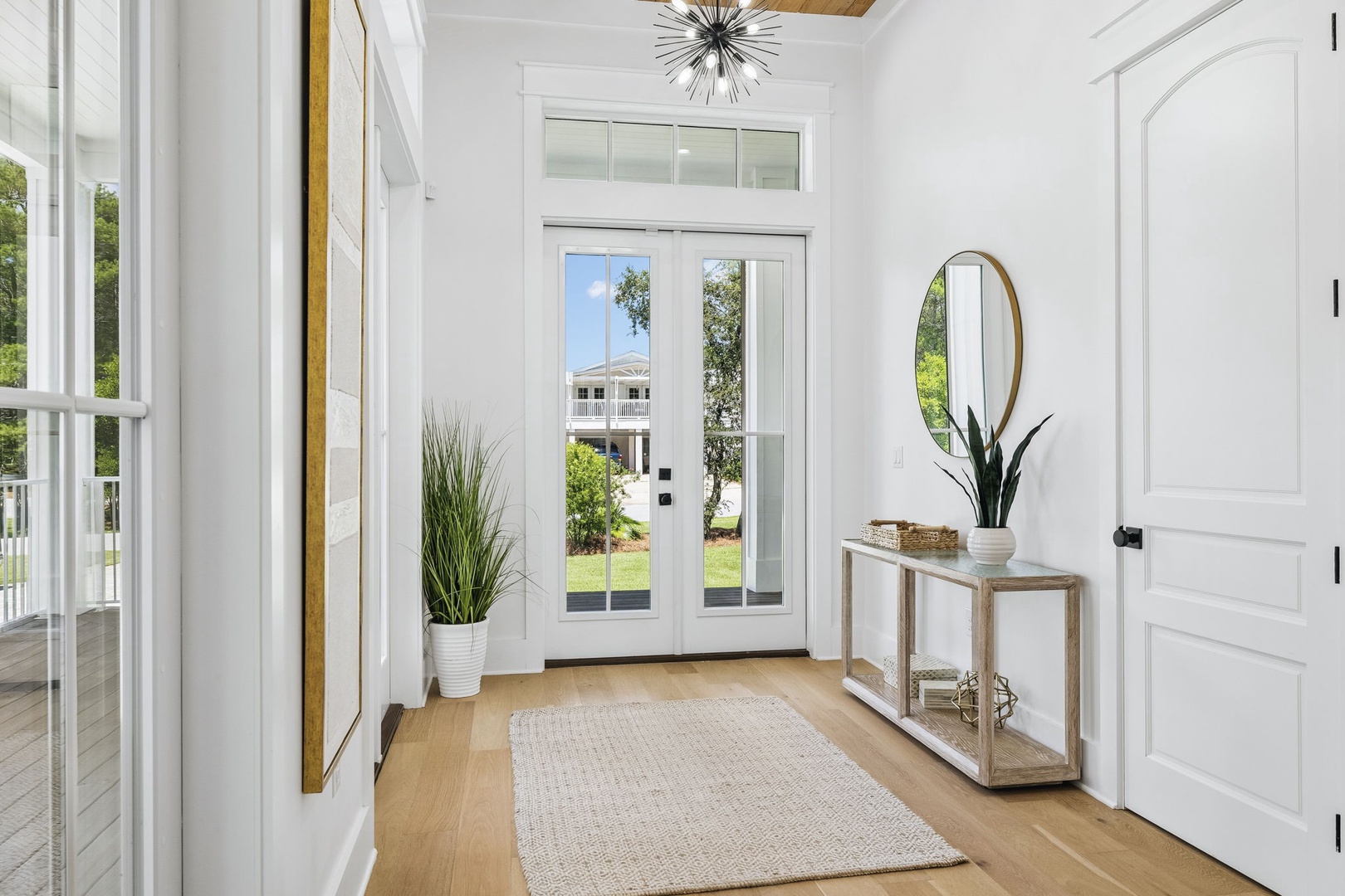 This bright entryway welcomes you with airy vaulted ceilings, warm wood floors, and a modern starburst chandelier. Sunlight streams through the glass doors, revealing the lush greenery beyond—your peaceful retreat awaits.