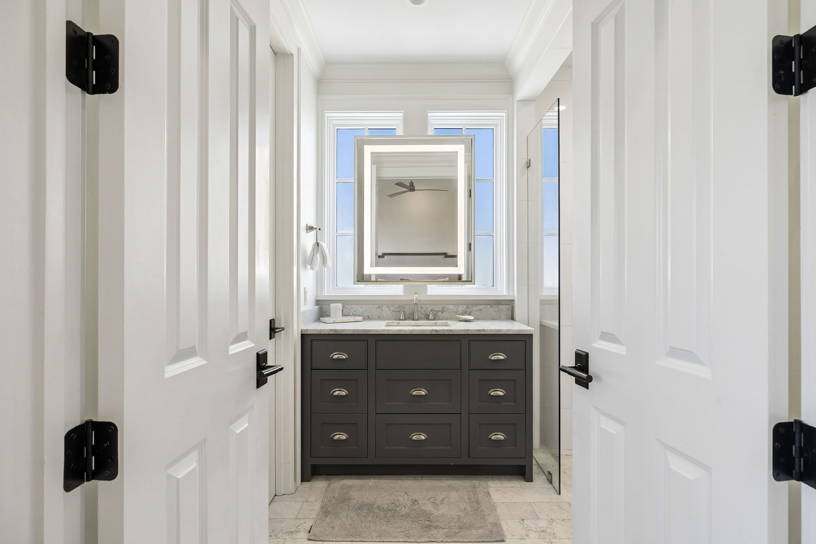Step into your private bathroom oasis. Classic white paneling frames a handsome gray vanity topped with marble, while natural light pours in through elegant windows, creating a fresh, calming retreat.