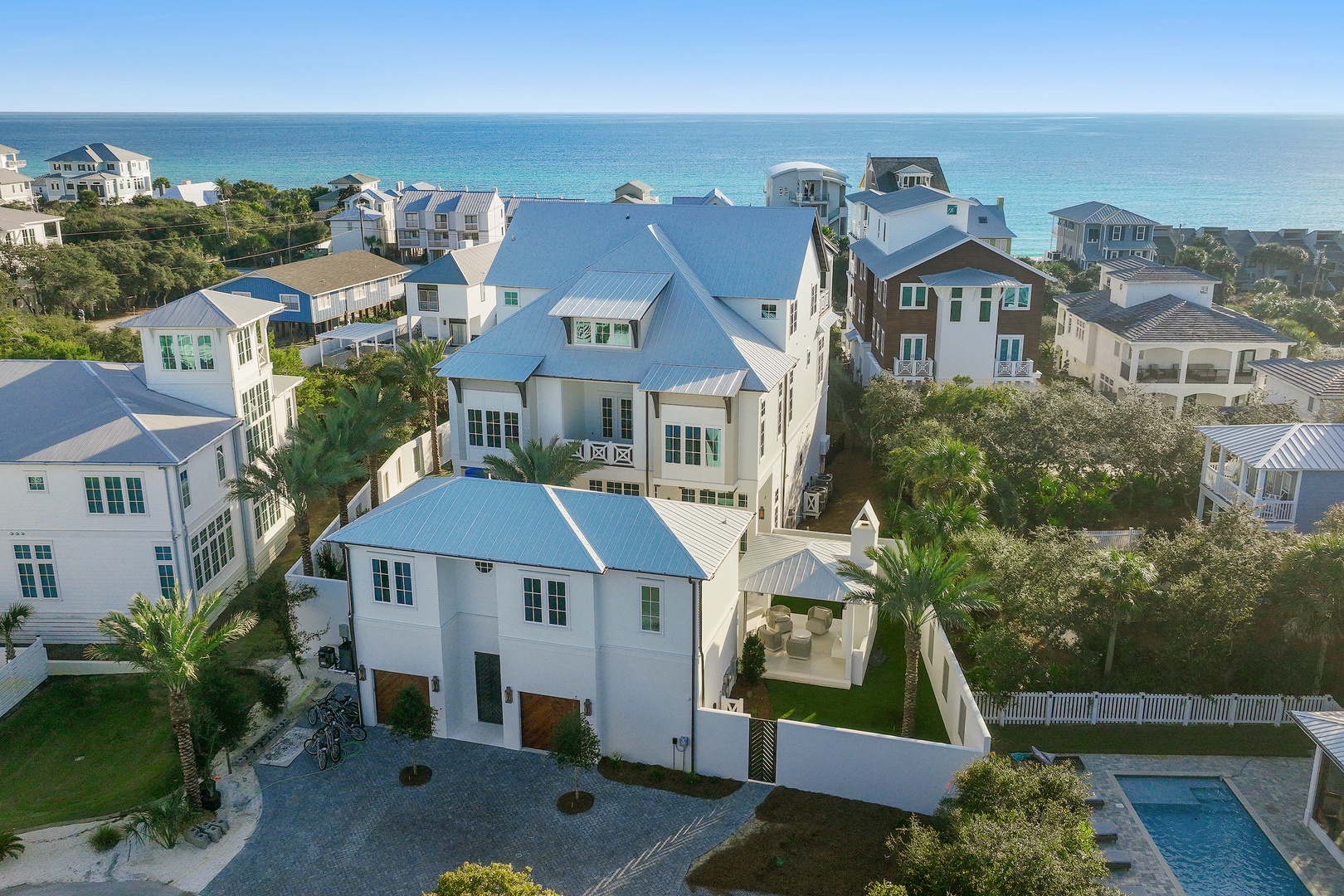 Aerial view of a spacious coastal property featuring blue metal roofing, palm trees, and a private pool, situated steps from turquoise Gulf waters in a serene beachside neighborhood.