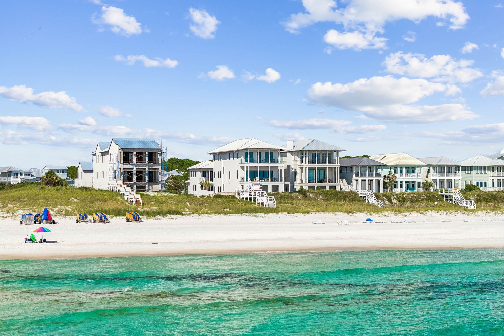A row of elegant beachfront homes lines the pristine white sand shore, with crystal-clear turquoise waters stretching out under a bright blue sky dotted with soft clouds.