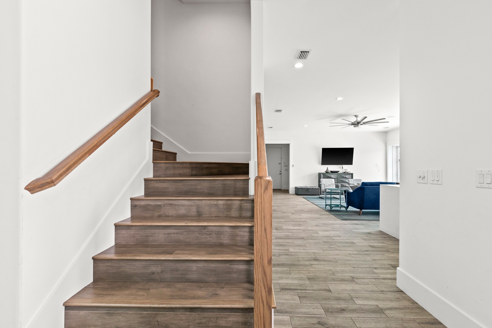 A bright, open stairway leads up to your living area, where modern comfort awaits with stylish seating, a mounted TV, and warm wood-toned floors throughout.