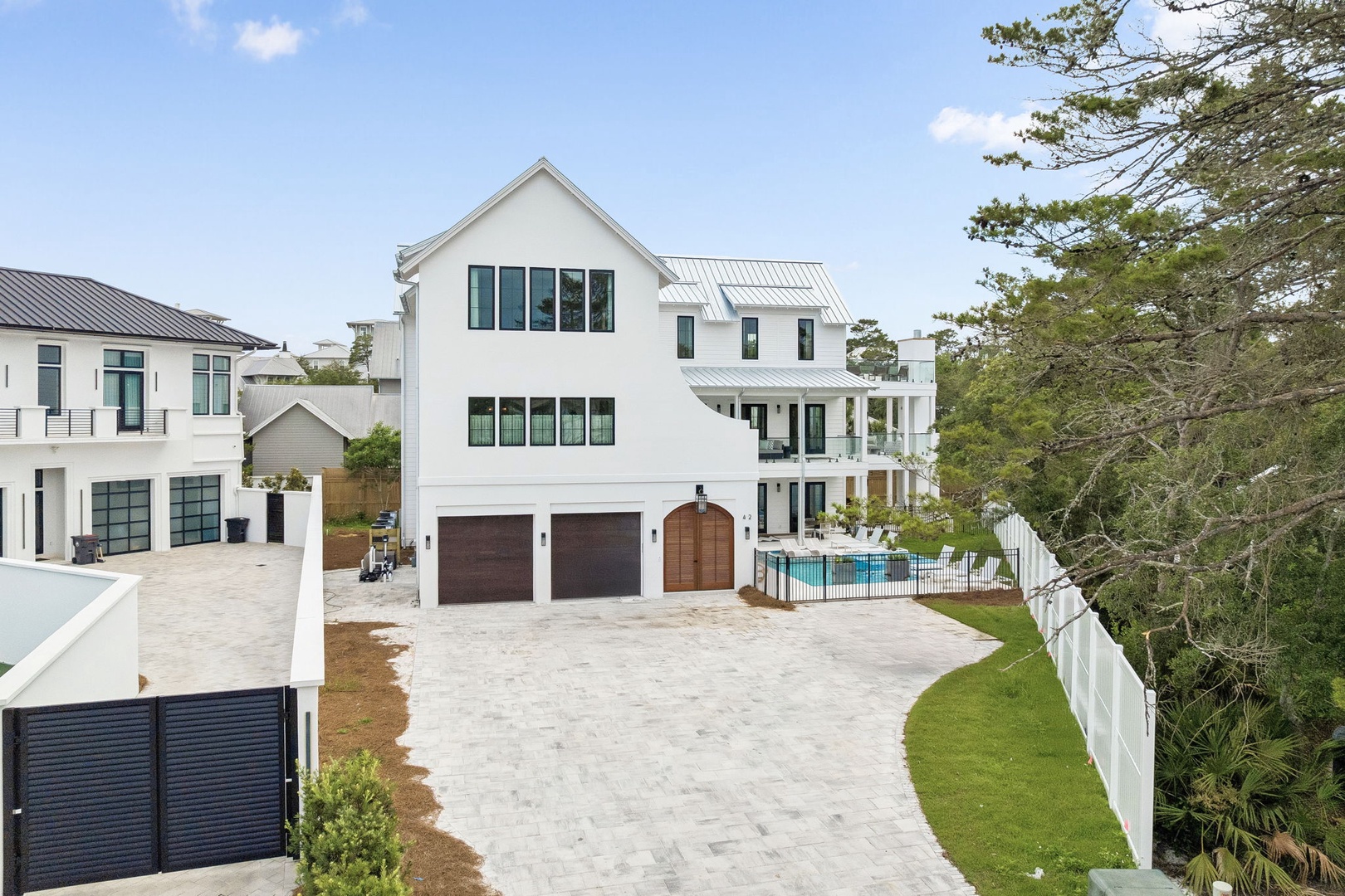 A modern coastal-style property featuring a pristine white exterior with a metal roof, spacious driveway, and private pool nestled in a residential setting near neighboring homes.