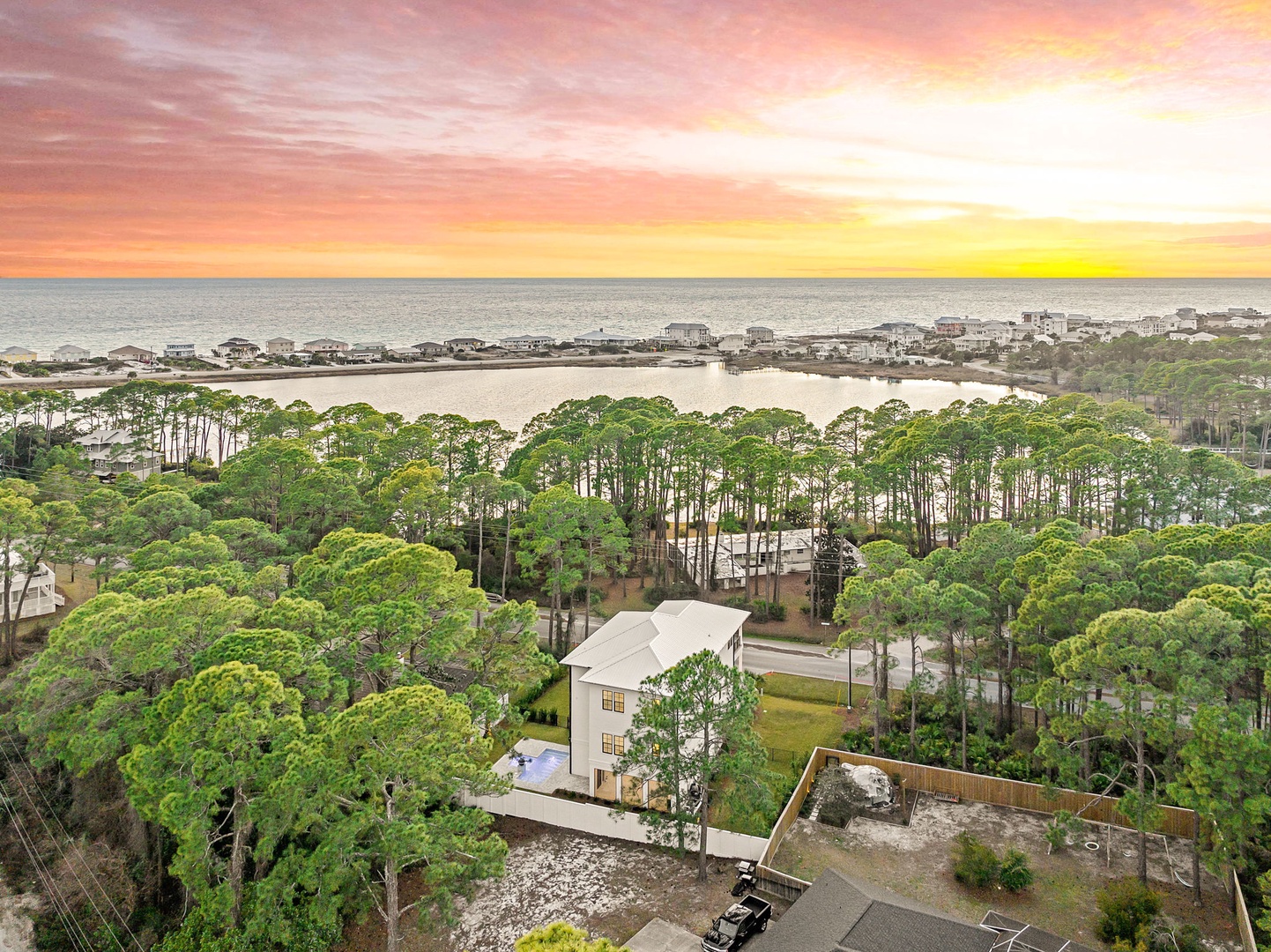 Aerial view showcasing the property nestled among lush coastal pines, with pristine beaches and dramatic sunset skies creating a picturesque Gulf Coast setting.