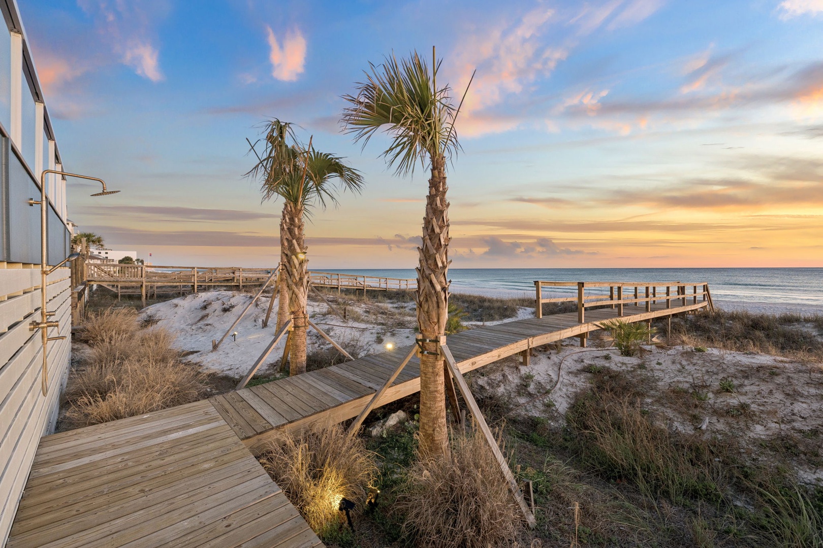 Wooden boardwalk winds through coastal dunes with palm trees, leading to pristine beach access during golden hour.