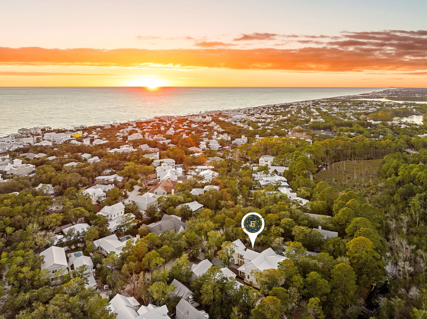 Aerial view of a coastal community nestled among lush trees, with the property location marked near the pristine shoreline at sunset.