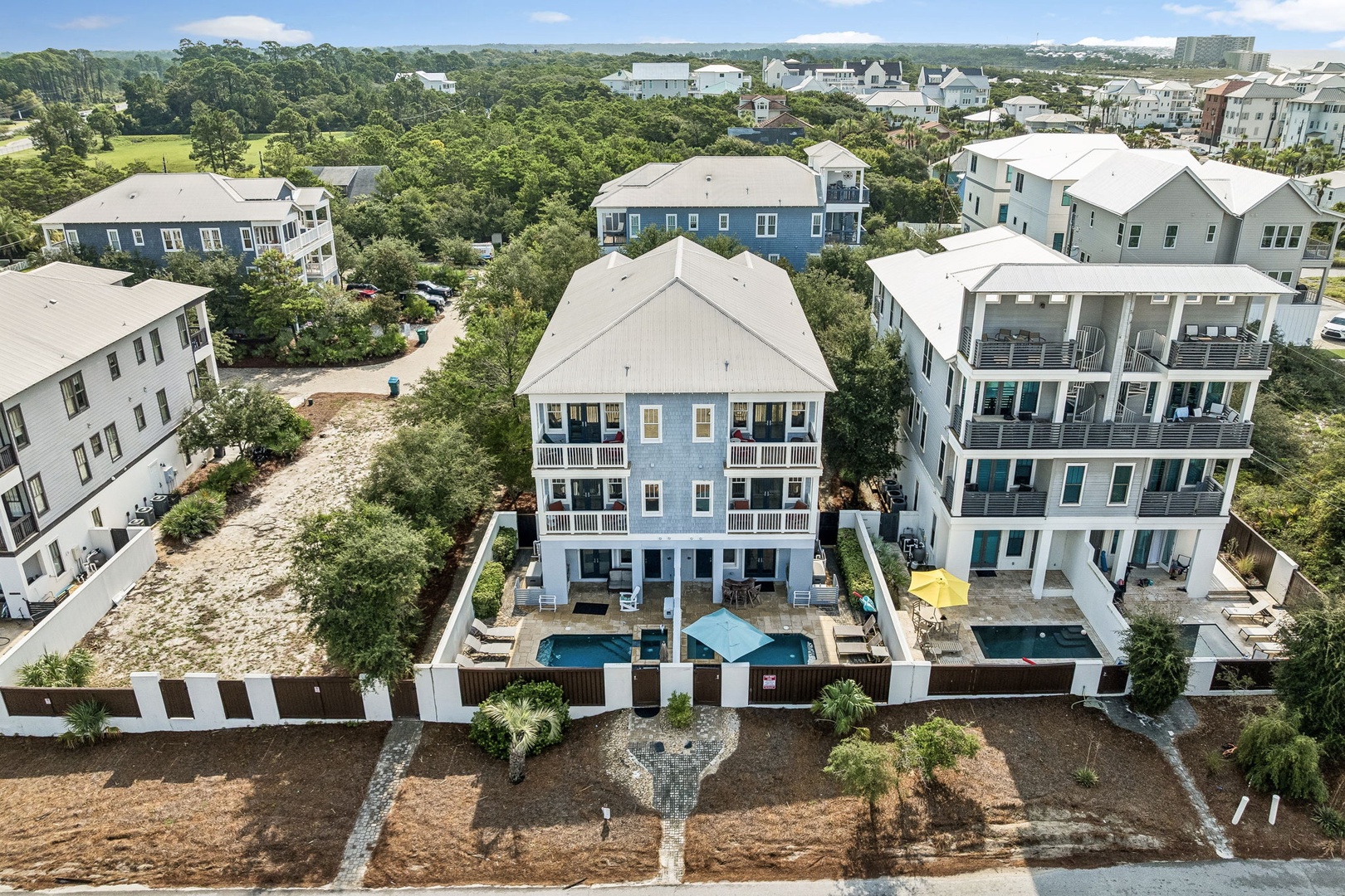 An aerial view reveals this coastal rental property nestled in a residential neighborhood surrounded by lush greenery and natural landscape, with multiple vacation homes sharing a communal area.
