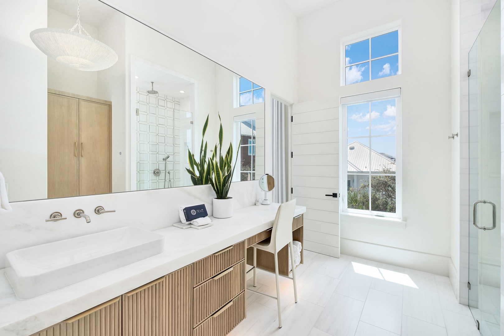Sunlight streams through tall windows, illuminating your serene bathroom retreat with its elegant double vanity, spa-like white tile shower, and warm natural wood accents.