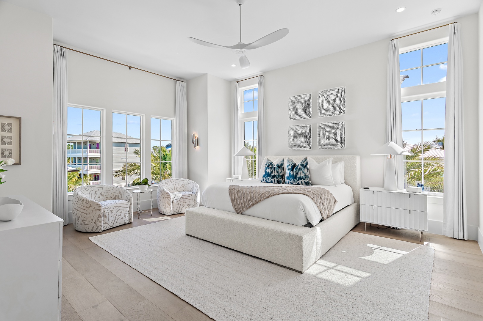 Unwind in this serene, sun-drenched bedroom with soaring ceilings, plush seating, and gentle breezes from a sleek ceiling fan. Large windows frame tropical palms and invite natural light to fill your peaceful retreat.
