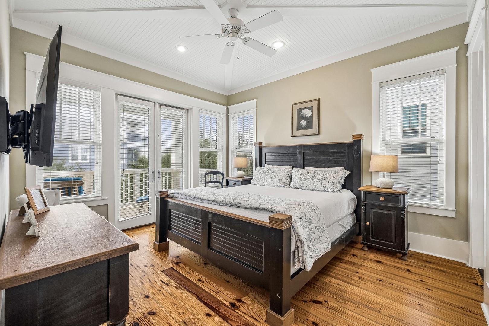 Unwind in your tranquil bedroom retreat, where natural light floods through multiple windows and a peaceful view awaits beyond the doors. The spacious room offers a plush king bed, vaulted ceilings, warm wood floors, and inviting coastal charm.