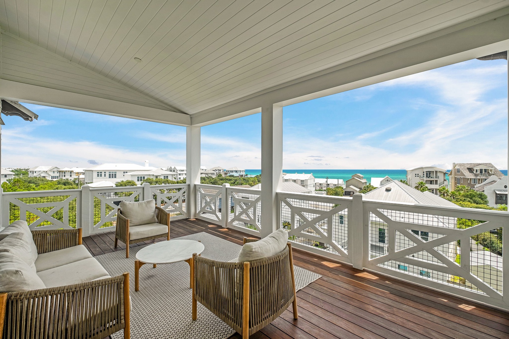 This covered porch overlooks the coastal neighborhood, with the turquoise waters and white sand beach visible in the distance beyond nearby homes and greenery.