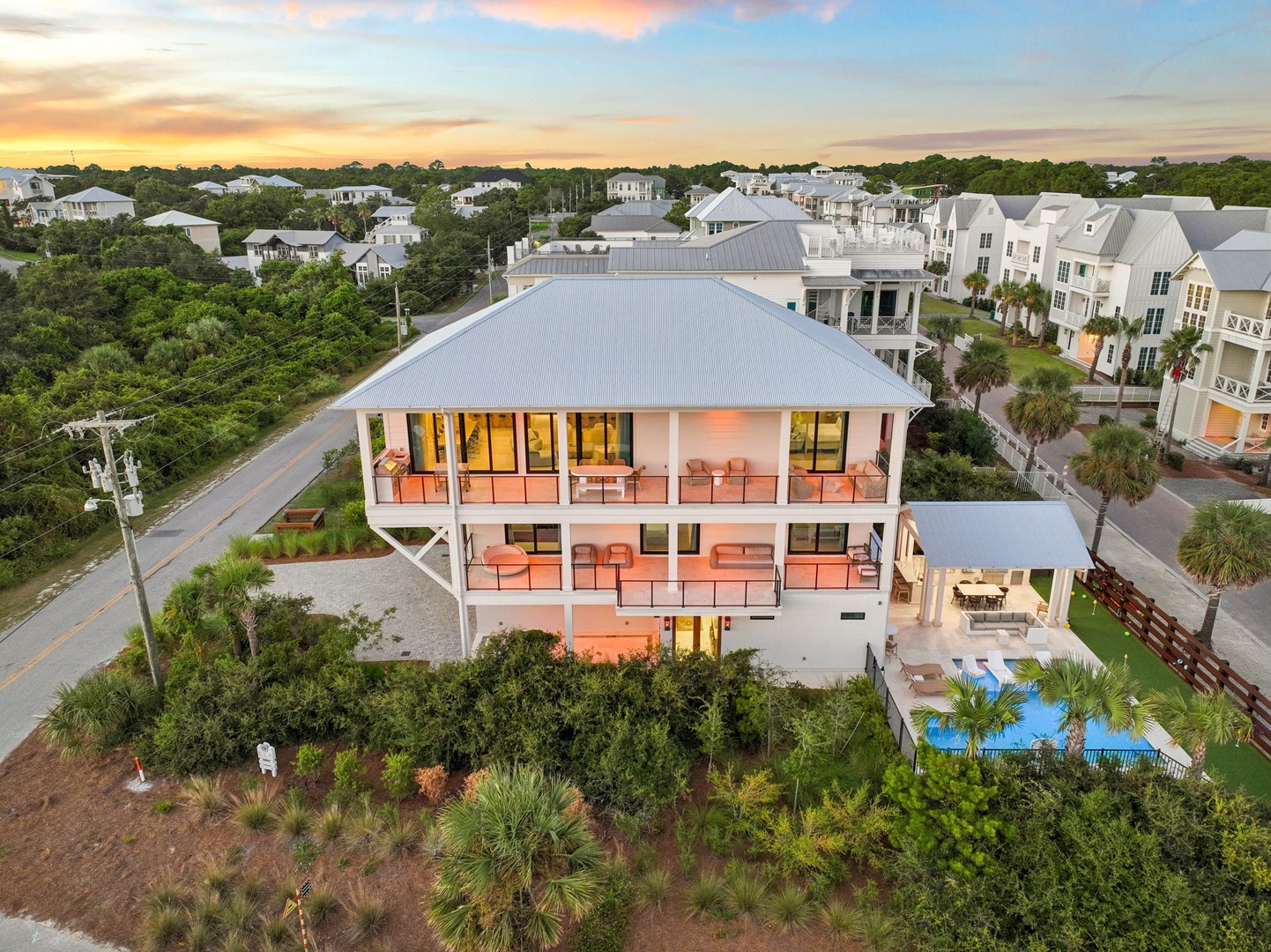 An aerial view captures this stunning coastal property nestled in a peaceful residential neighborhood. The multi-level home features spacious balconies and a private pool, surrounded by lush tropical landscaping and neighboring beach homes.