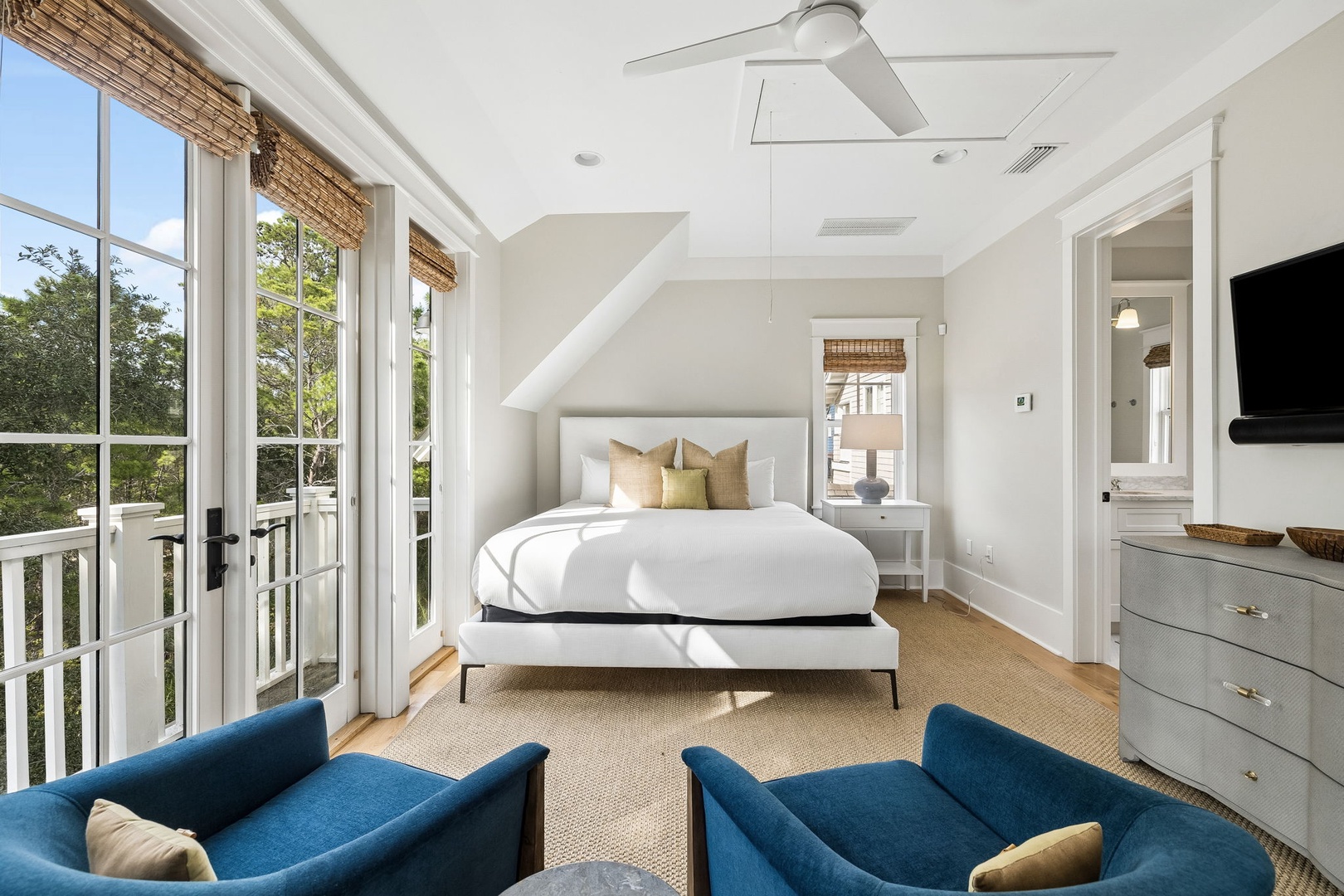 Your bright, airy retreat features a plush bed dressed in crisp white linens and accent pillows. Relax in cozy seating while natural light streams through French doors opening to your private balcony, framed by leafy treetop views.