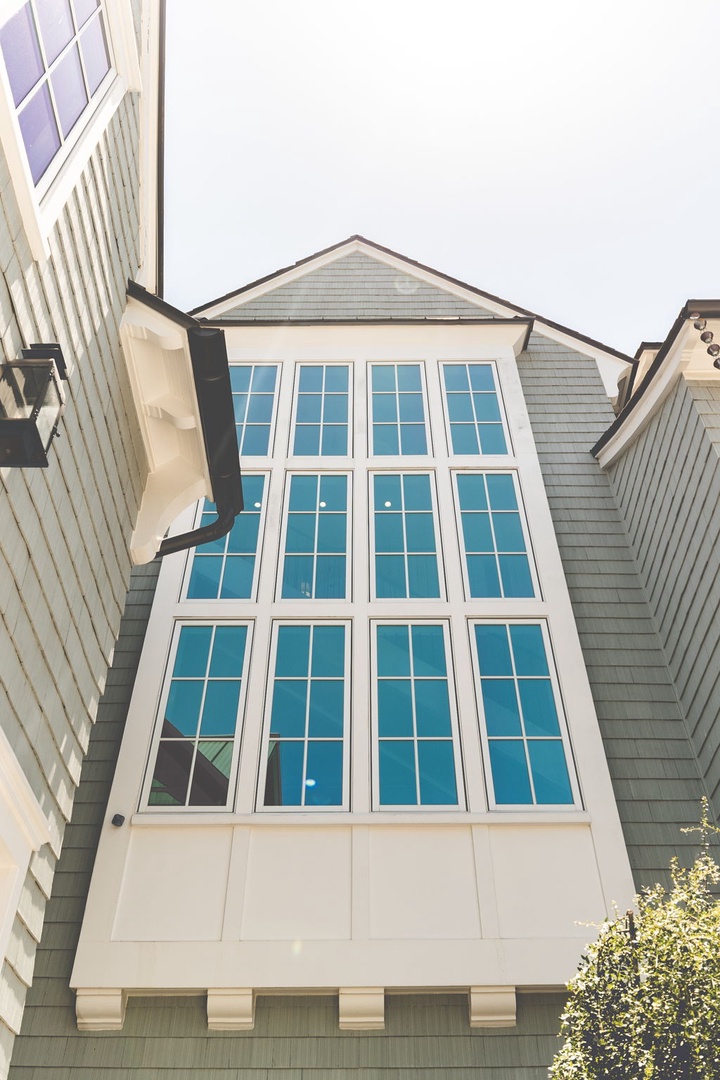 A modern coastal-style property exterior with gray shingle siding, white trim, and a striking three-story wall of windows that frames the entrance and surrounding residential area.