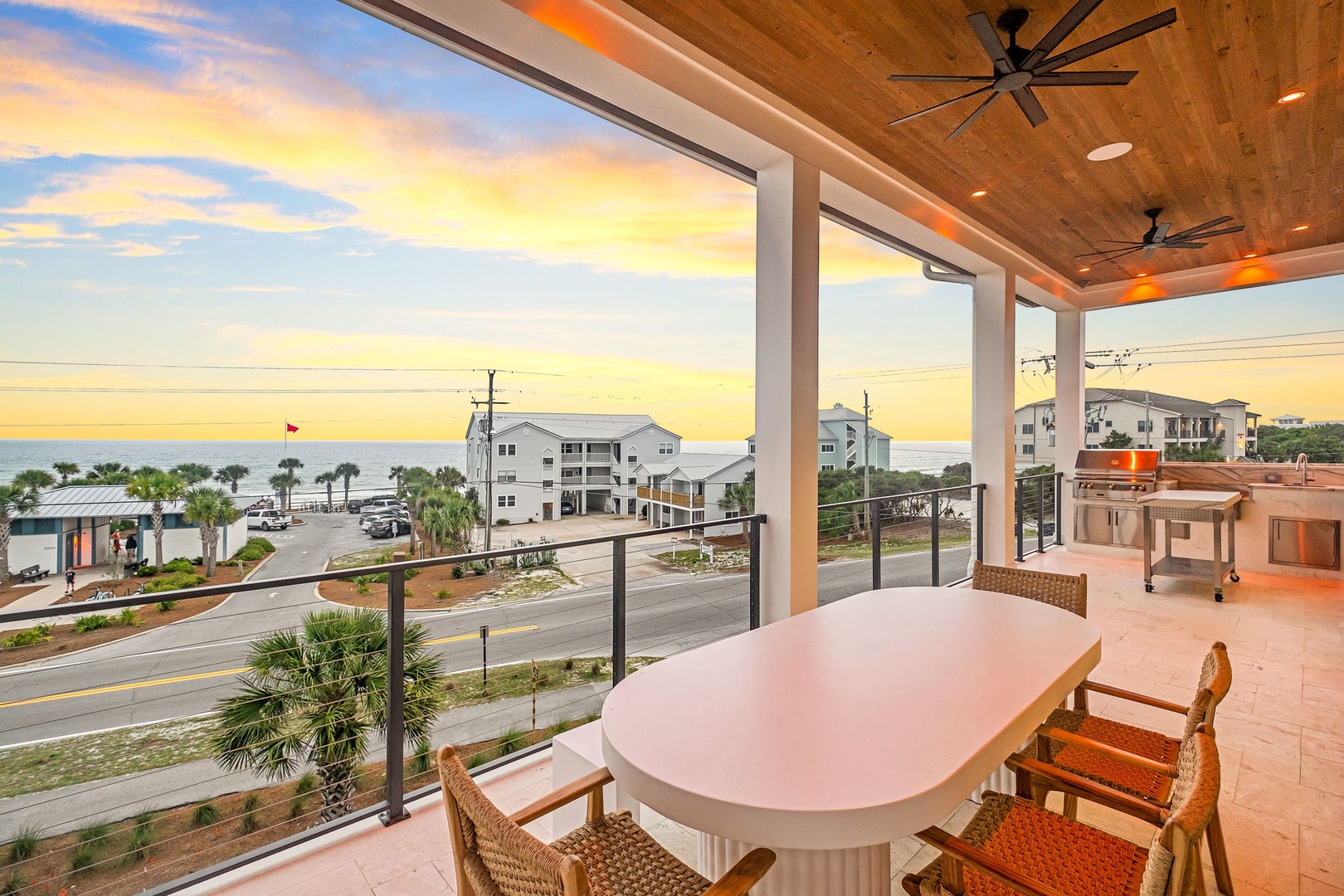 A covered terrace with stunning sunset views framing palm trees, nearby buildings, and the shimmering sea beyond. Features dining table, outdoor kitchen, and ceiling fans.