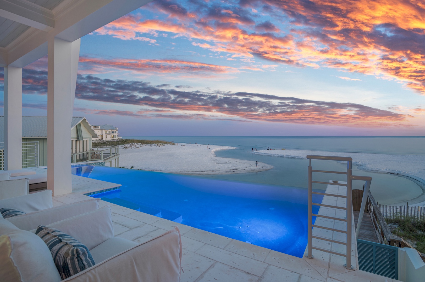 A stunning coastal property featuring an infinity pool overlooking pristine beach and ocean waters, with neighboring beachfront homes dotting the sandy shoreline—framed by dramatic evening skies.