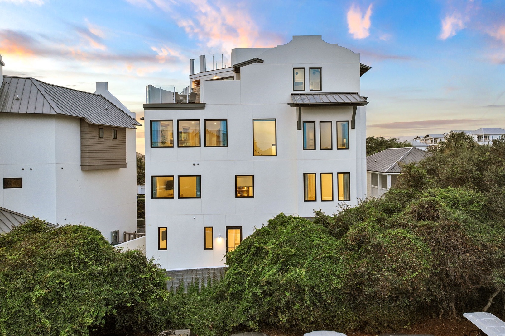 Modern coastal architecture nestled among lush greenery in a peaceful beachside neighborhood, with warm interior lighting creating an inviting evening ambiance.