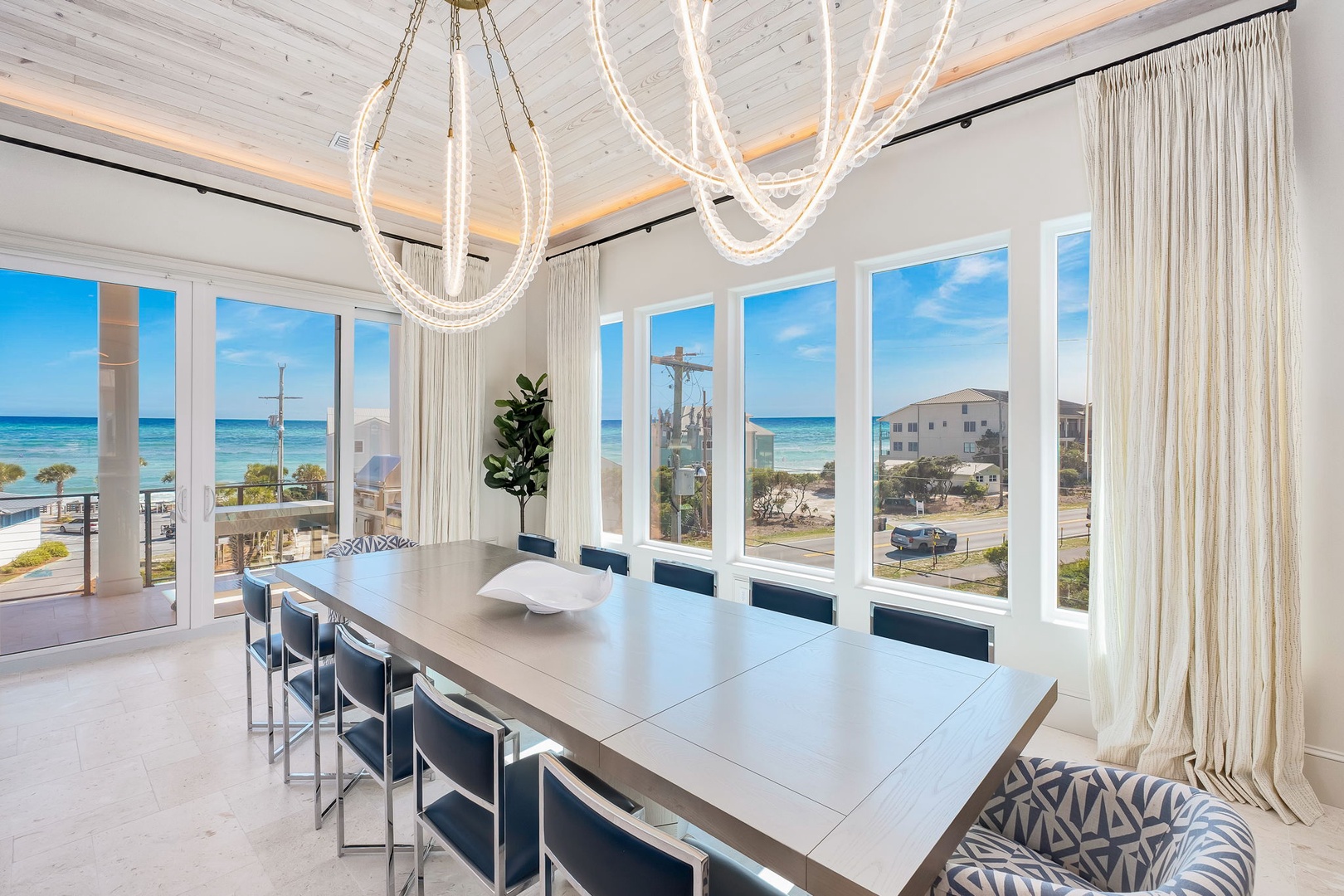 A coastal property featuring expansive ocean views through floor-to-ceiling windows, surrounded by beachside vegetation and neighboring vacation homes.