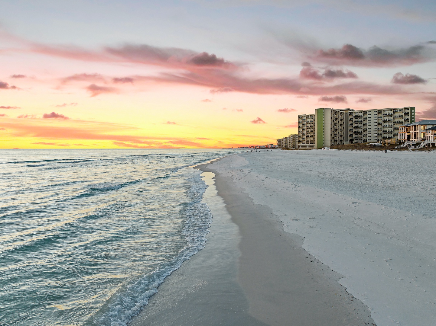 Stunning beachfront location with pristine white sand and gentle waves just steps from modern coastal buildings at sunset.
