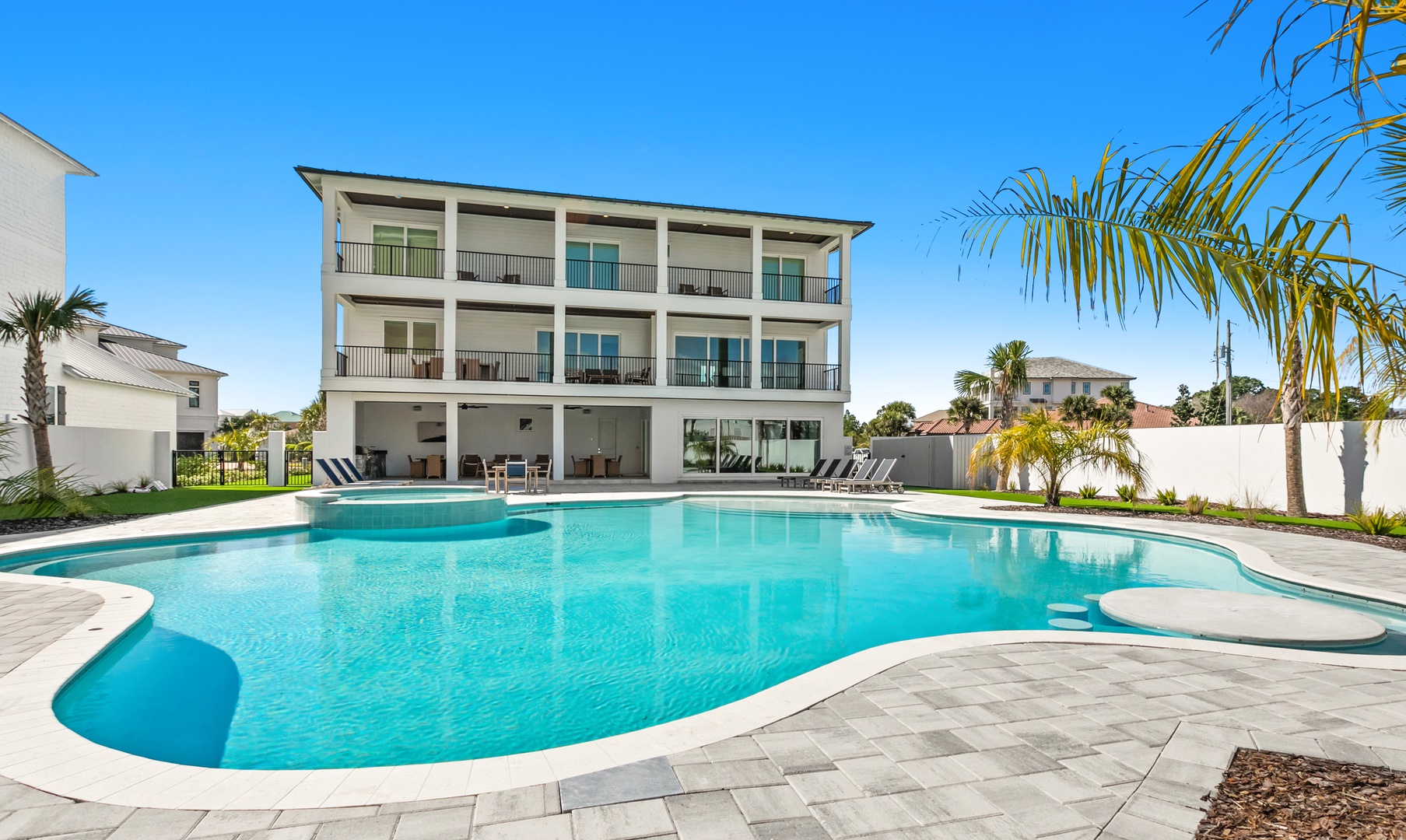 A spacious modern coastal residence features multiple private balconies overlooking an expansive curved swimming pool, surrounded by tropical landscaping and paved pool deck with lounging areas.