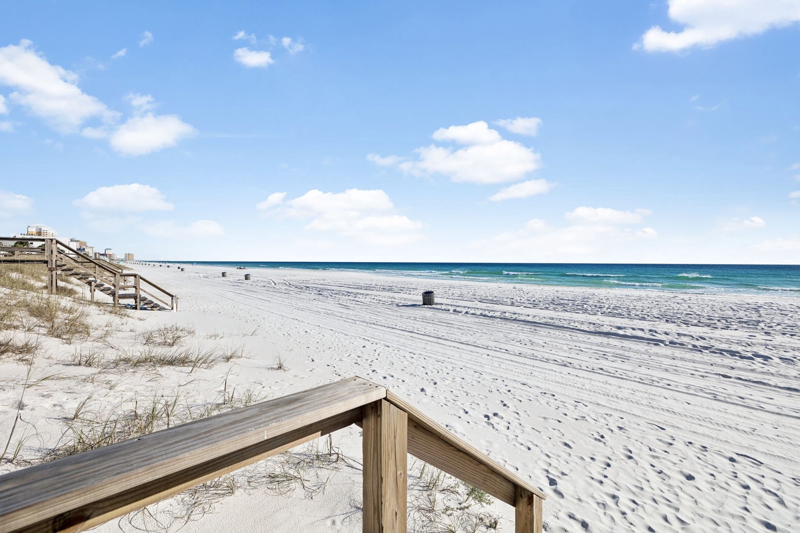 Pristine white sand beach stretches endlessly under blue skies, with wooden walkways providing easy access to the Gulf waters.