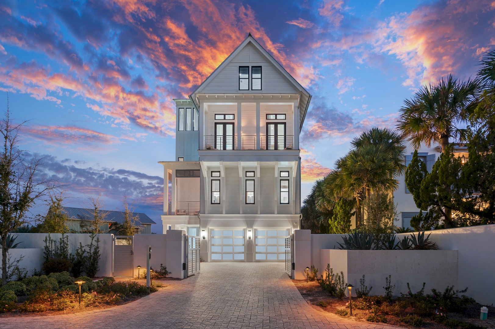 A stunning three-story beach home framed by palm trees and a dramatic sunset, welcoming guests at the end of a paver driveway. The surrounding landscaped courtyard and coastal architecture set the stage for an unforgettable stay.