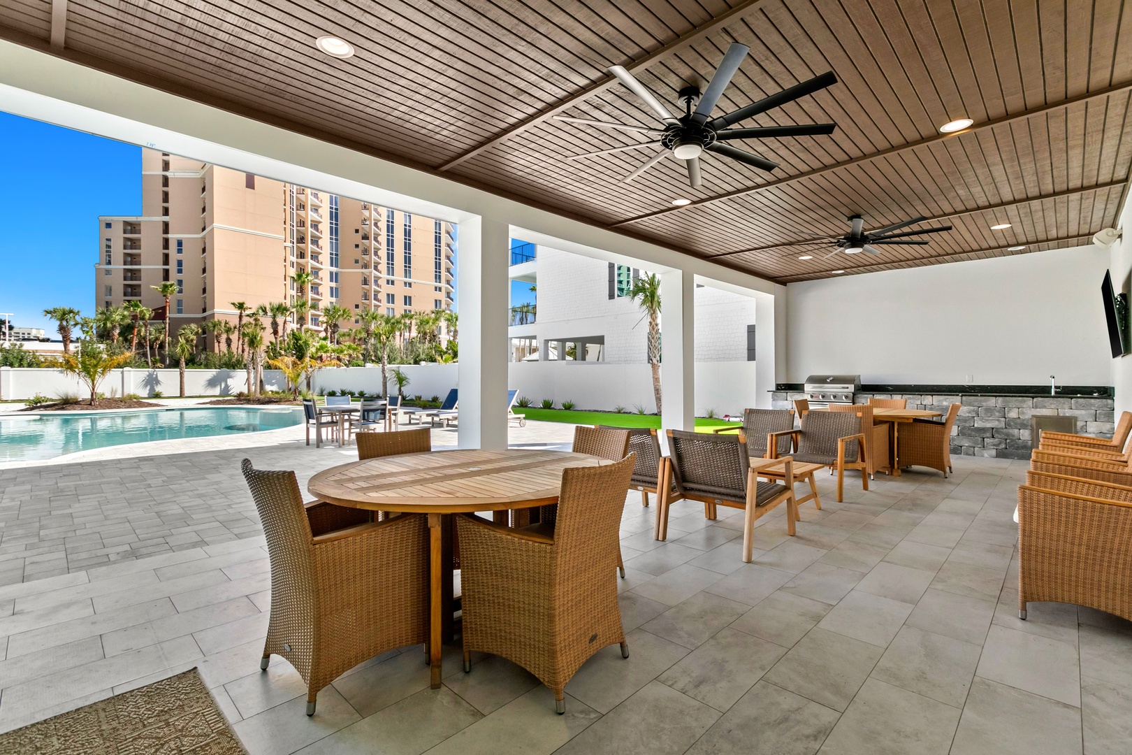 A poolside haven with a built-in grill and comfortable seating under ceiling fans invites you to unwind. Palm trees and modern urban towers frame the horizon, blending relaxation with city energy.