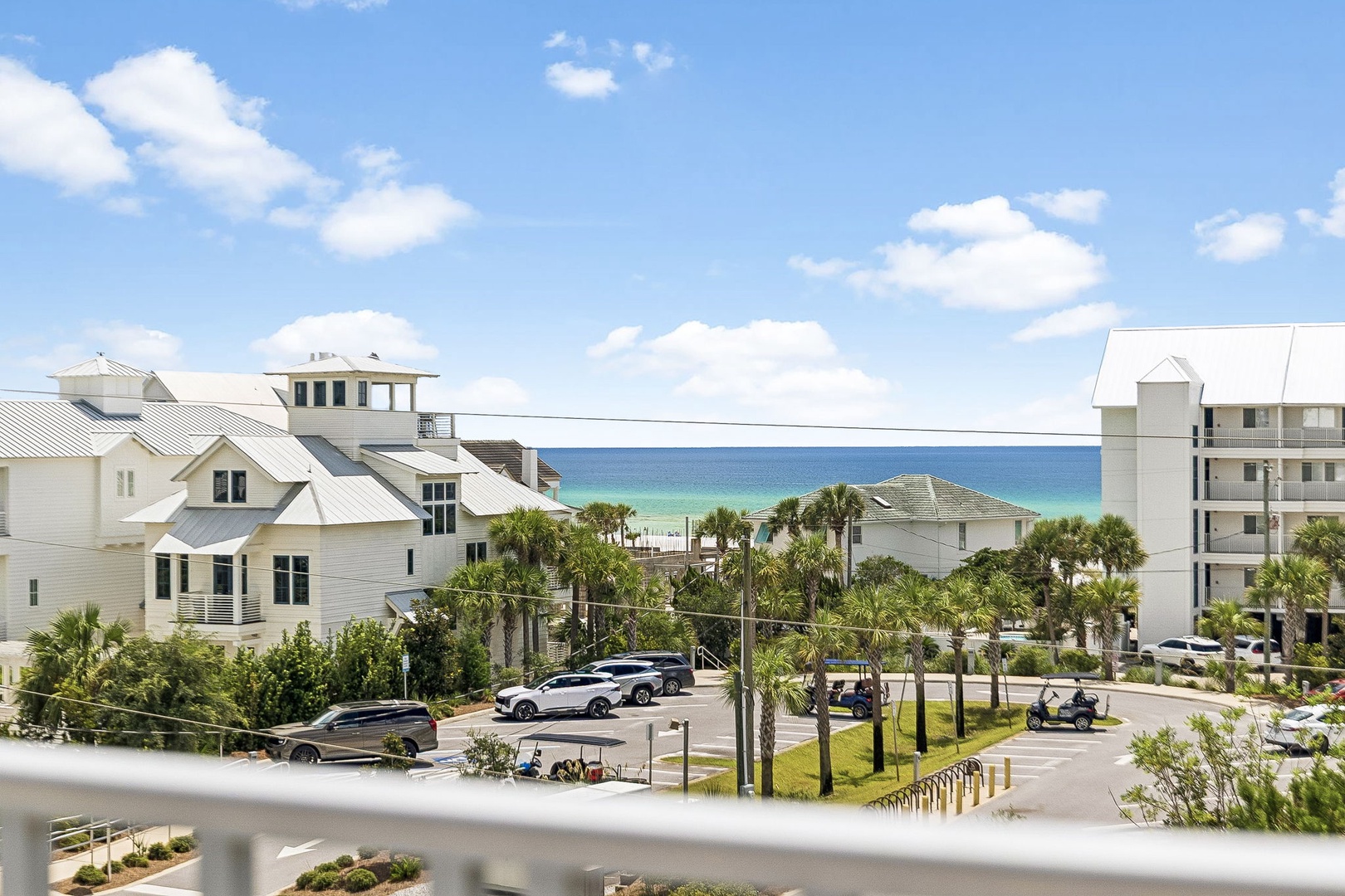 The coastal property sits in a serene beach neighborhood beneath clear blue skies, with palm trees dotting the surrounding streets and the inviting ocean visible just beyond the homes.