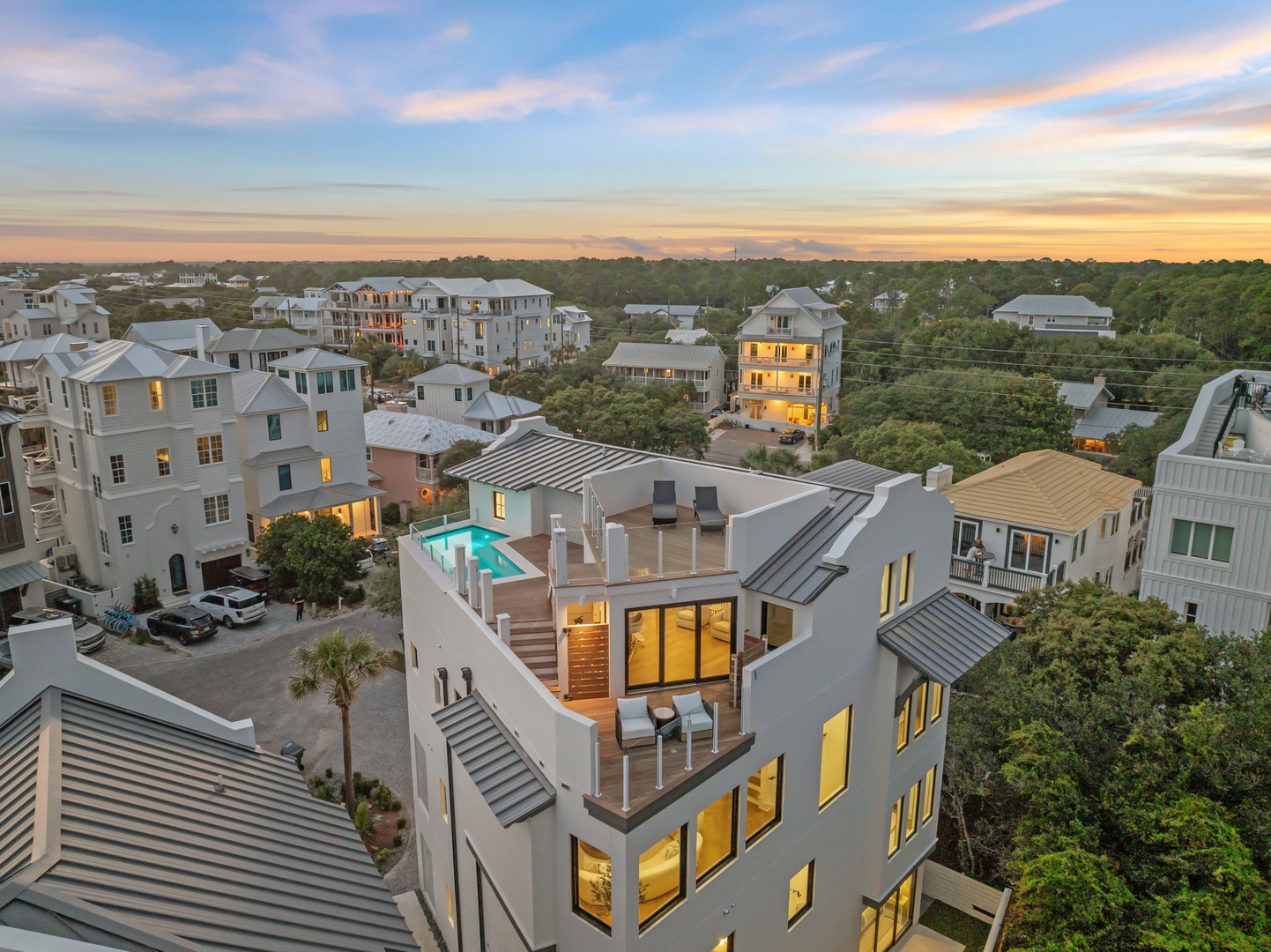 Aerial view of a modern vacation home with a rooftop deck, swimming pool, and multiple outdoor terraces, set within a residential coastal community at sunset.
