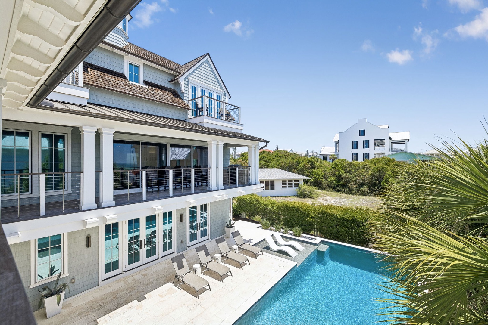 Multi-level coastal property featuring an inviting pool deck with lounge chairs, surrounded by lush vegetation and neighboring beach homes under a bright blue sky.