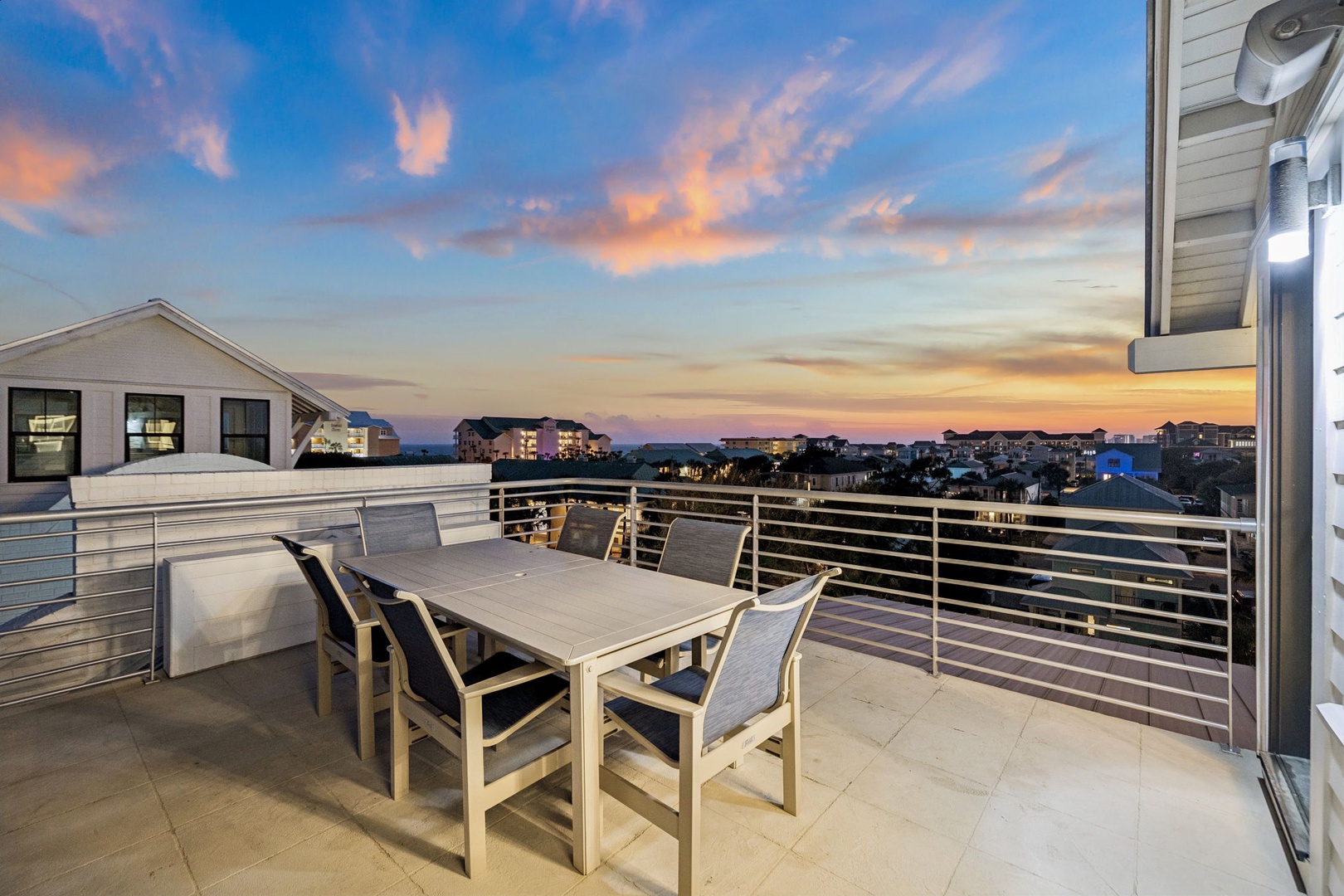 Spacious rooftop terrace with dining table overlooking coastal neighborhood under colorful sunset sky.