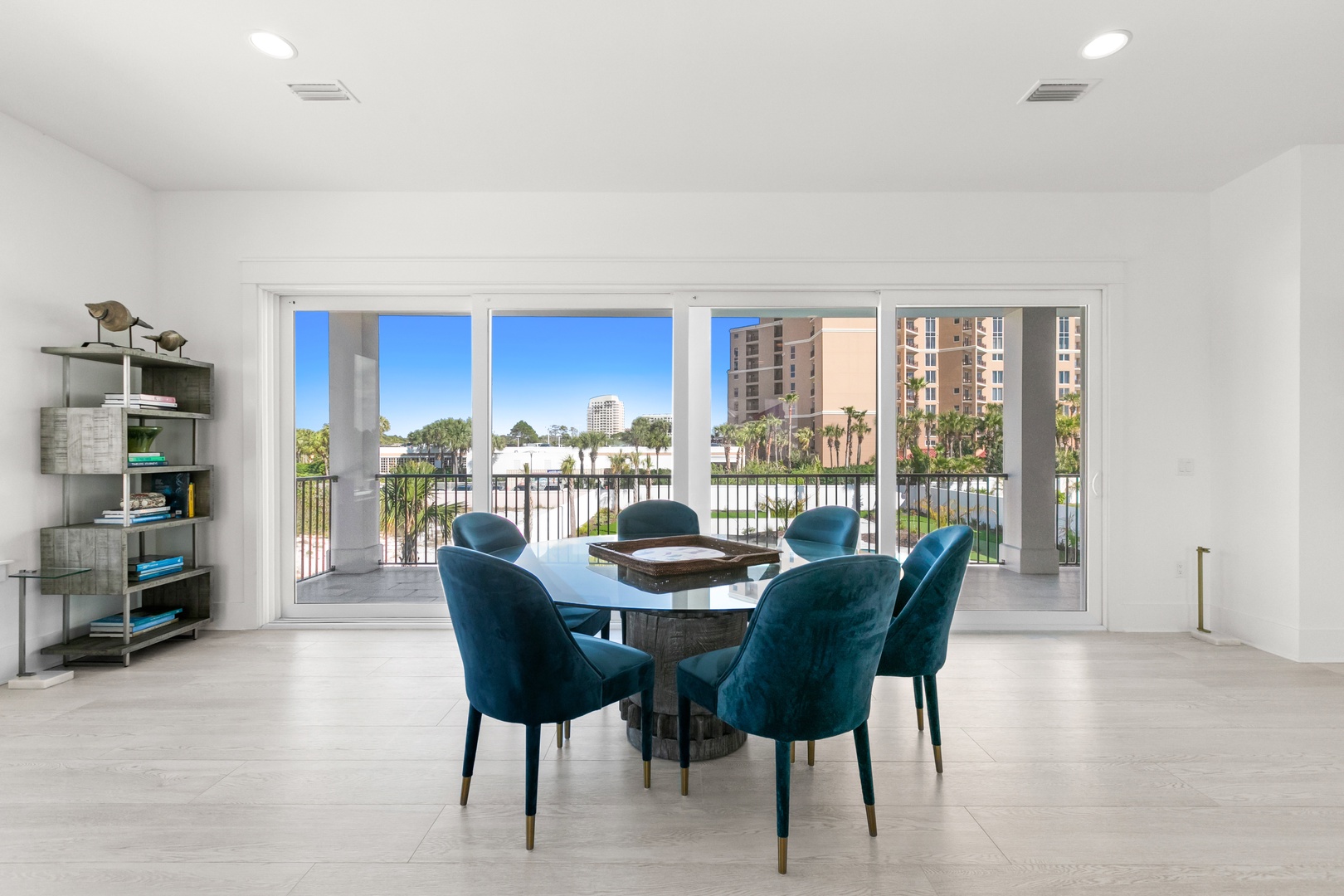 Step into your bright, open dining space where plush teal chairs await shared meals. Floor-to-ceiling windows frame the waterfront beyond, filling your room with natural light and fresh coastal air.