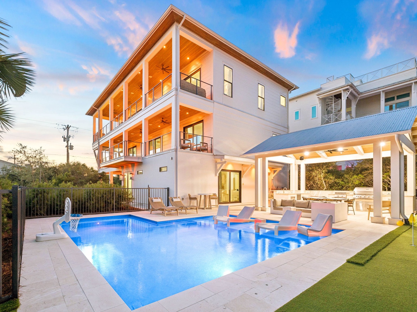 Modern three-story coastal retreat with private pool and spacious covered patio, illuminated by warm lighting as day transitions to dusk. Surrounded by lush greenery in a peaceful neighborhood.
