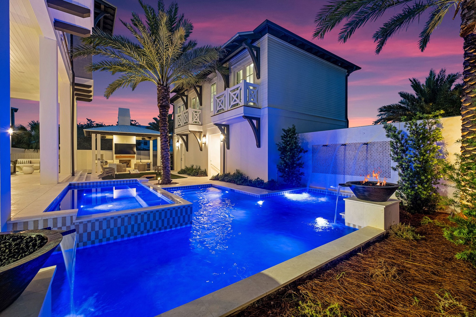 A stunning luxury property illuminated at twilight, featuring a pristine pool area surrounded by palm trees and tropical landscaping under a painted sky.