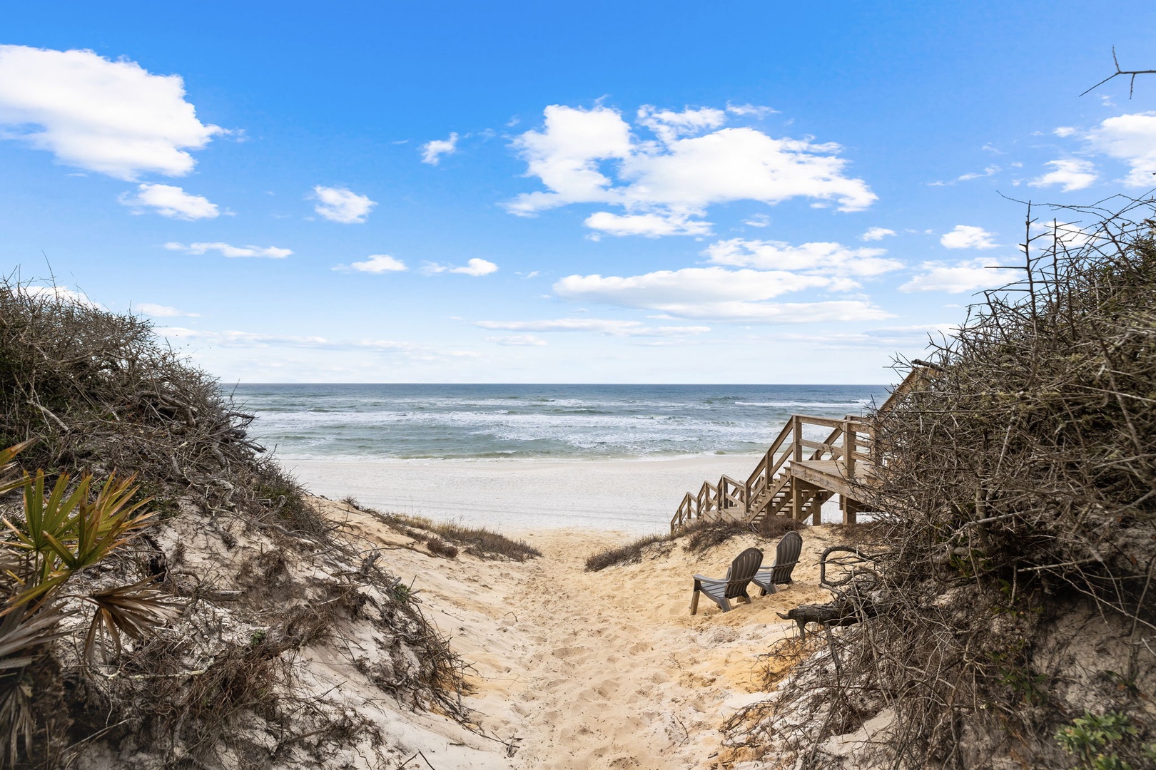 The property features a private sandy beach path with ocean views, framed by natural dunes and a charming wooden stairway. Inviting chairs on the sand offer a peaceful spot to soak in the coastal beauty.
