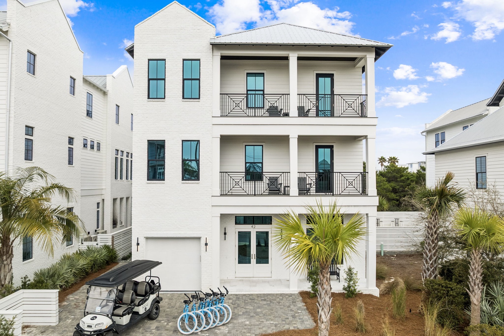 Modern three-story coastal home with double balconies, surrounded by tropical palms in an upscale beach community.
