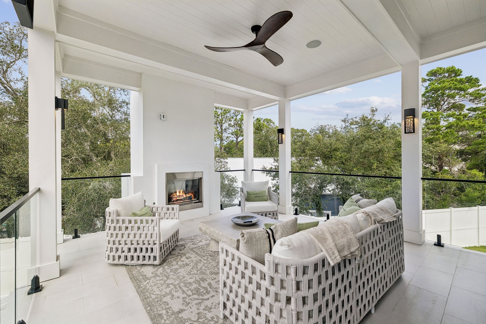 A covered outdoor terrace with elegant woven seating and a cozy fireplace, surrounded by leafy treetop views—your perfect spot to unwind under the ceiling fan.