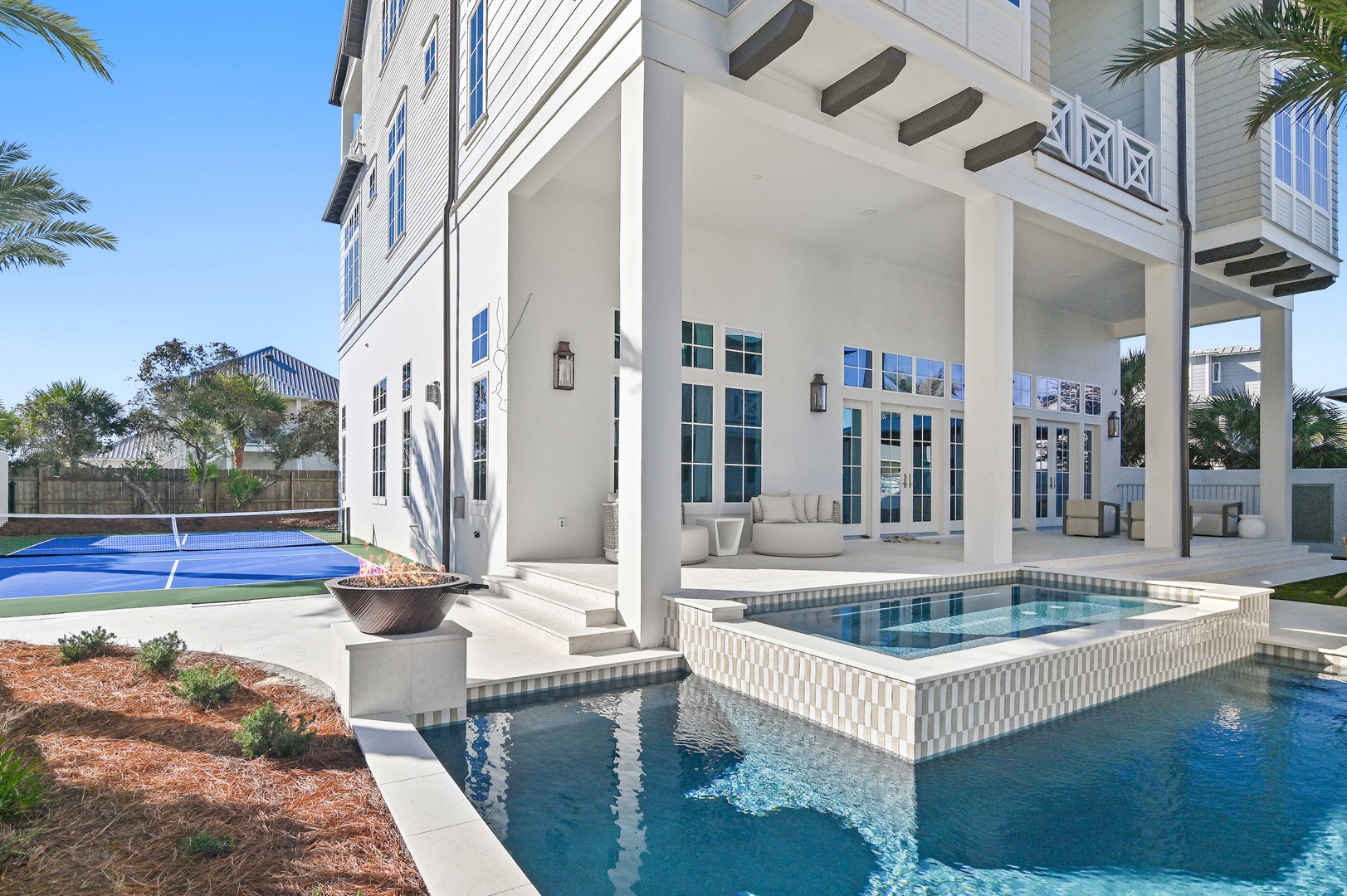 A luxurious coastal property showcasing elegant architecture with pristine swimming pool, spa, tennis court, and lush palm-lined grounds under sunny blue skies.