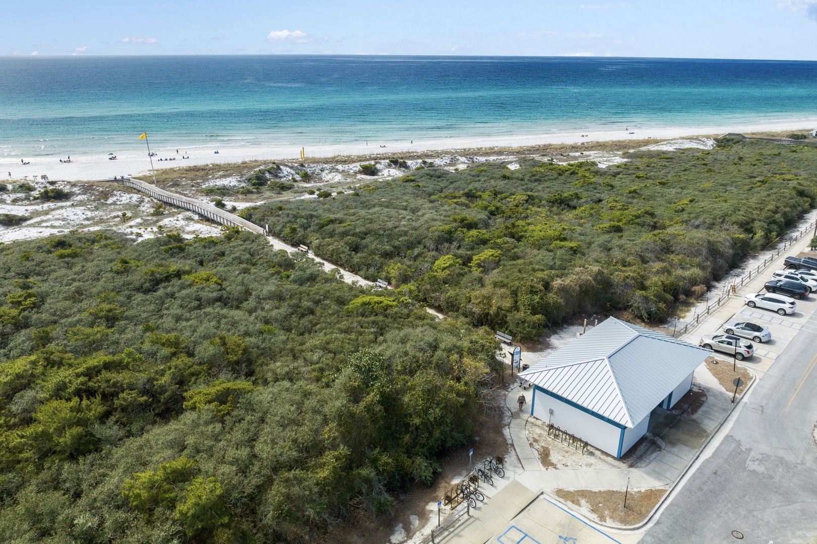 Aerial view showcasing pristine coastal setting with turquoise waters, sandy beach, natural dunes, and convenient parking near beachfront facilities.