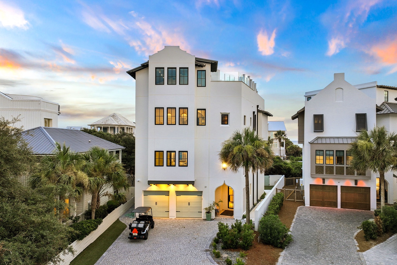A striking white coastal property surrounded by palm trees and neighboring homes, photographed during golden hour as the sky displays soft pink and blue hues.
